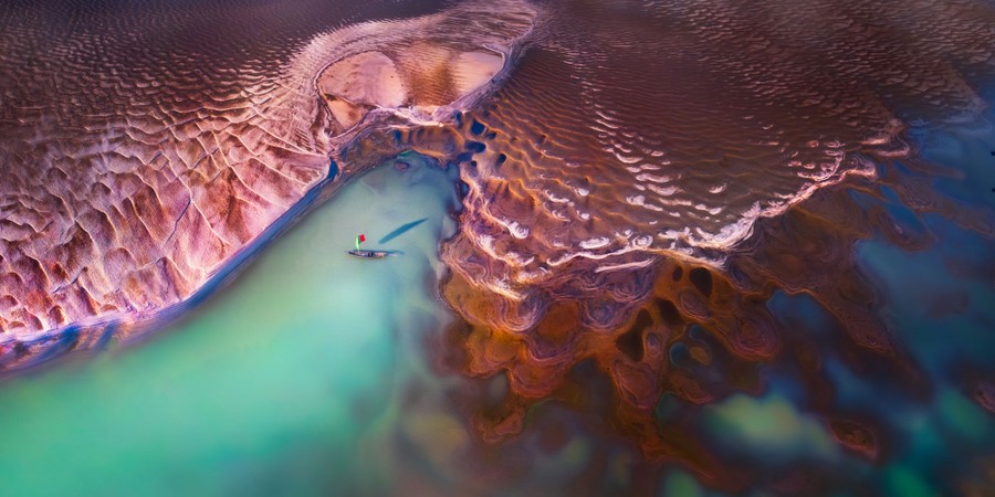 A small sailboat sits anchored along a layered sandy river bank.