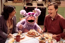 two people sitting at dinner table laughing next to a purple puppet