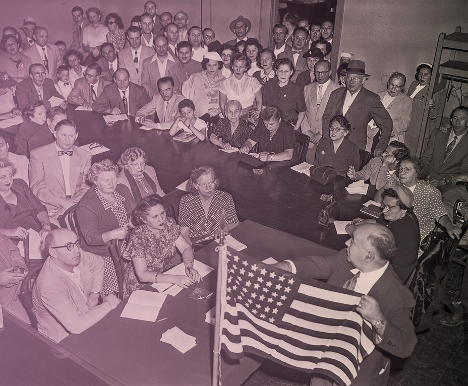 archival photo of room full of people sitting at tables paying attention to man lecturing and pointing to American flag