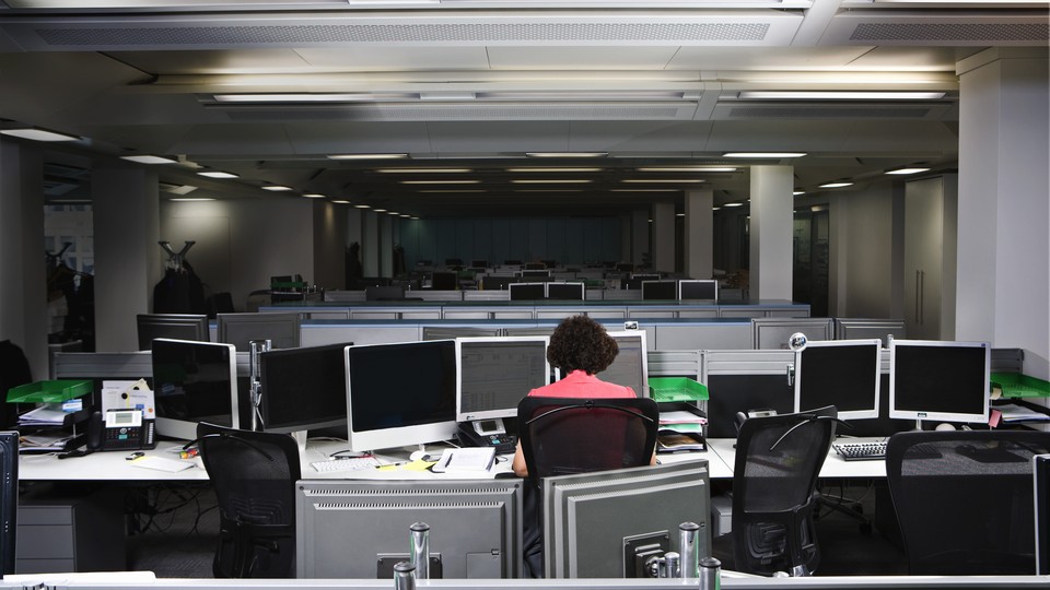 A worker sits alone in a dimly lit office, surrounded by computer monitors