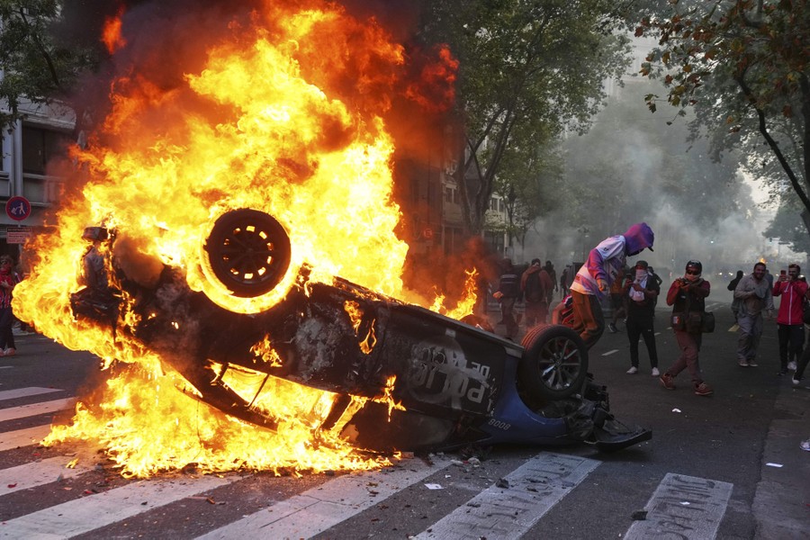 An overturned police car burns in a street as nearby protesters look on.