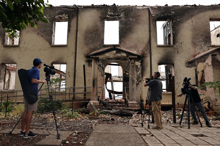 Several journalists operate cameras on tripods outside of a fire-destroyed building.