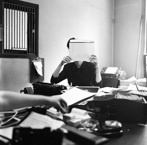 Black and white photo of a person working at a cluttered desk