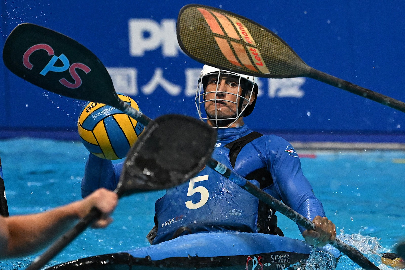 An athlete wearing a helmet while sitting in a canoe in a swimming pool leans back to throw a ball while being defended against by other others with paddles.