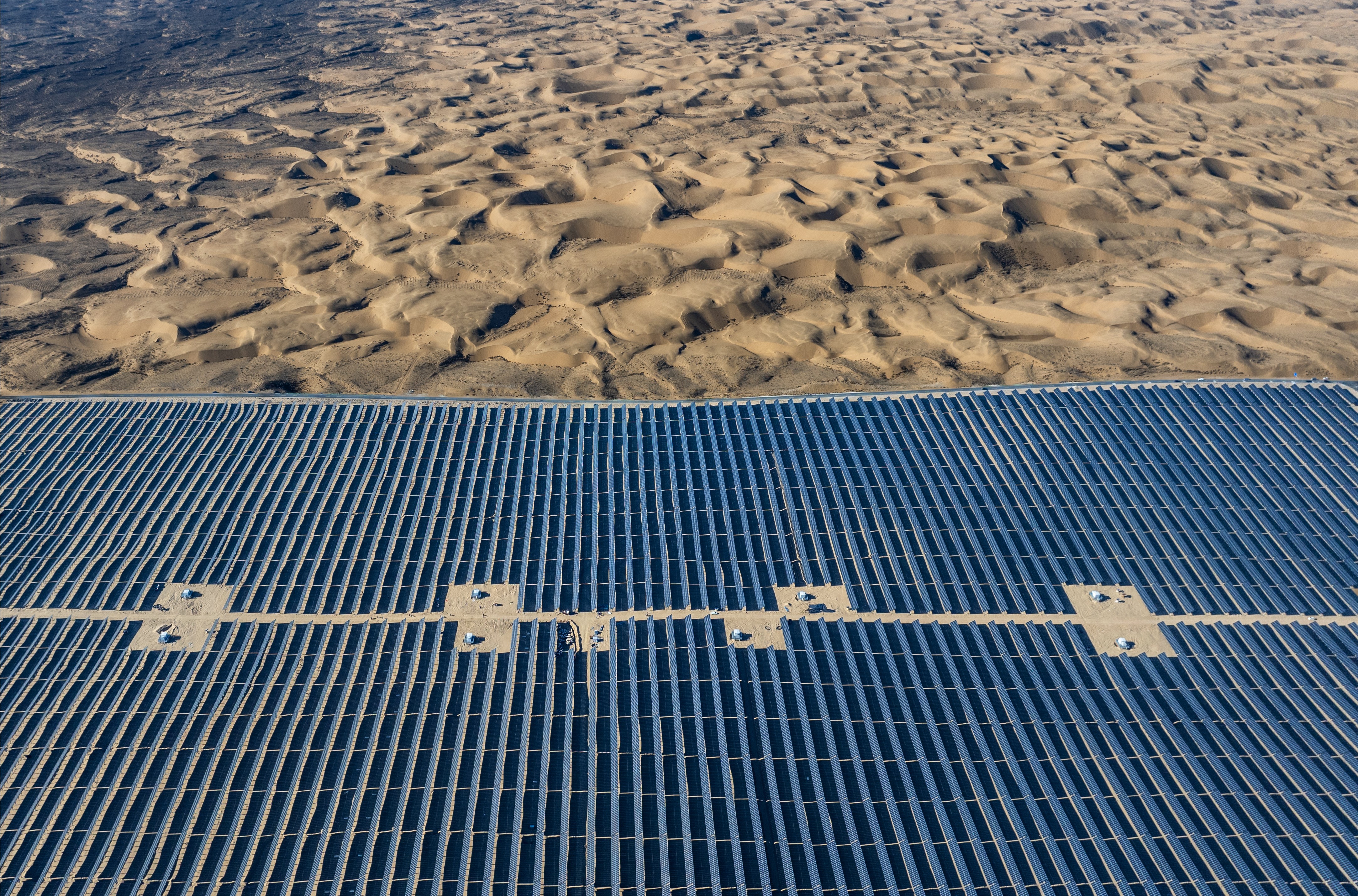Many solar panels stretch across a desert, with dunes visible beyond.