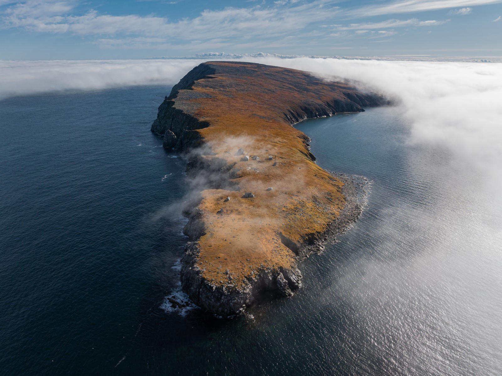 An aerial view of a group of small abandoned structures on a treeless island with steep cliff shorelines