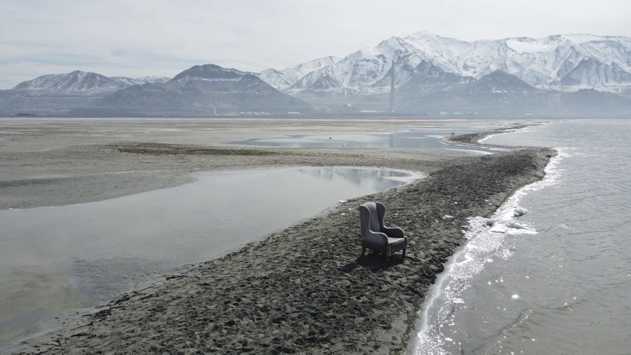 An upholstered chair sits alone on a narrow sandbar along the Great Salt Lake in front of a mountain range.