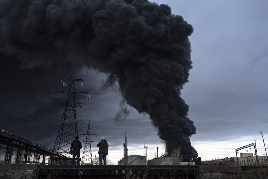 Two people watch a column of black smoke rise above industrial buildings.