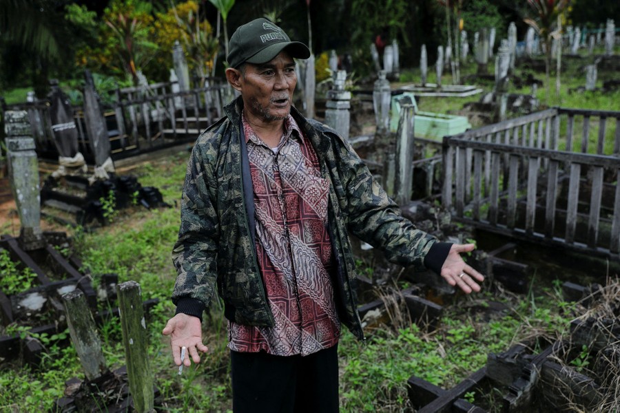 A man stands in a cemetery, gesturing toward the graves.