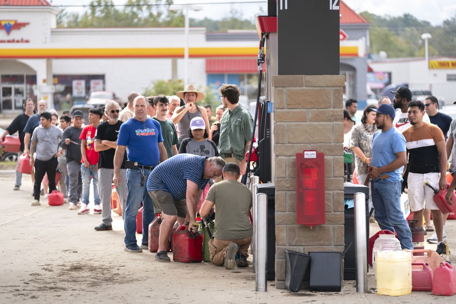Dozens of people stand in line near a gas-station pump, waiting to fill plastic jugs with gasoline.