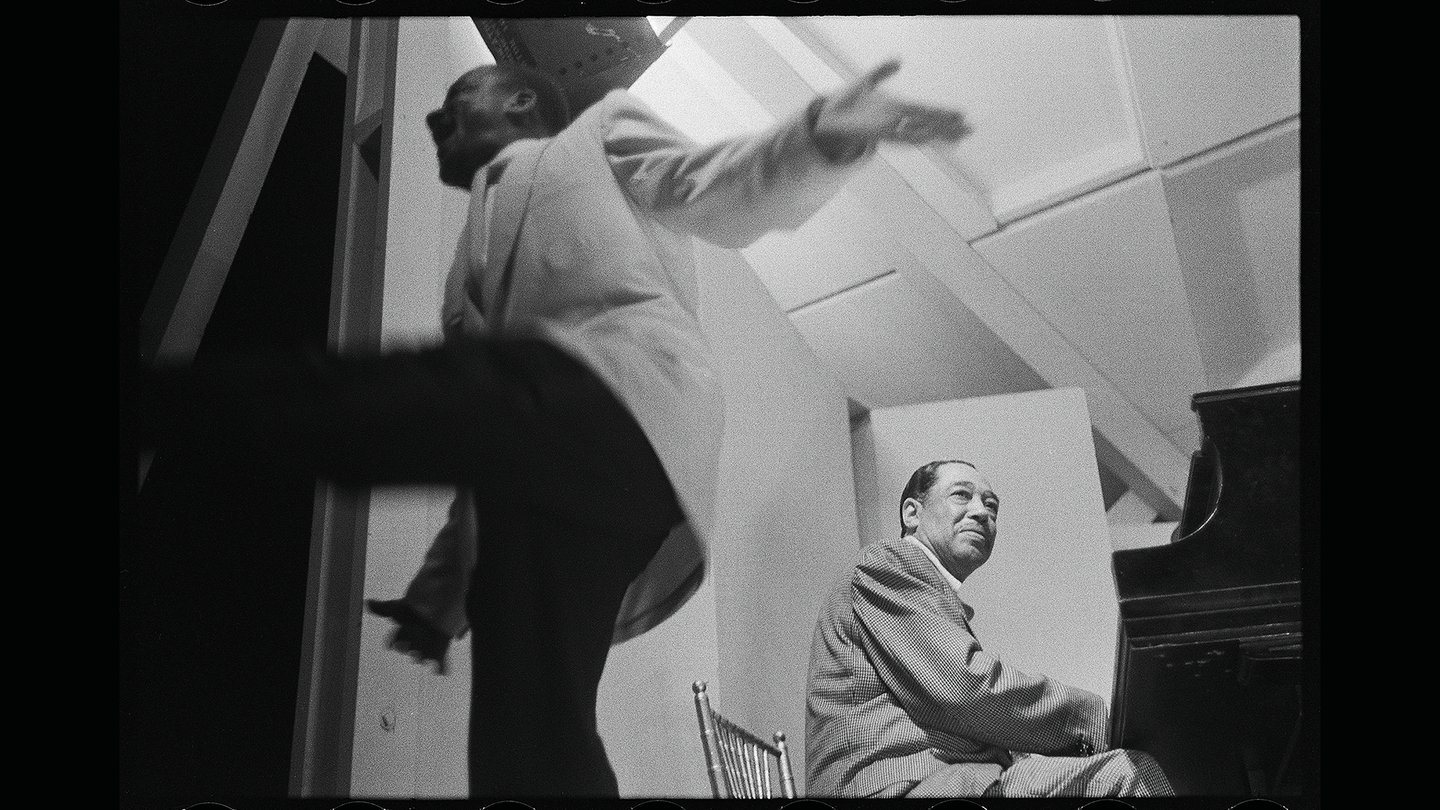 black-and-white photo of two men in suits taken from low angle, one dancing with arms out and one sitting on chair playing piano