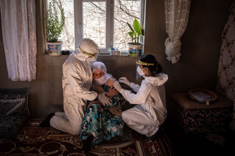 A pair of nurses hold and vaccinate an older woman who is seated on the floor in a room in a house.