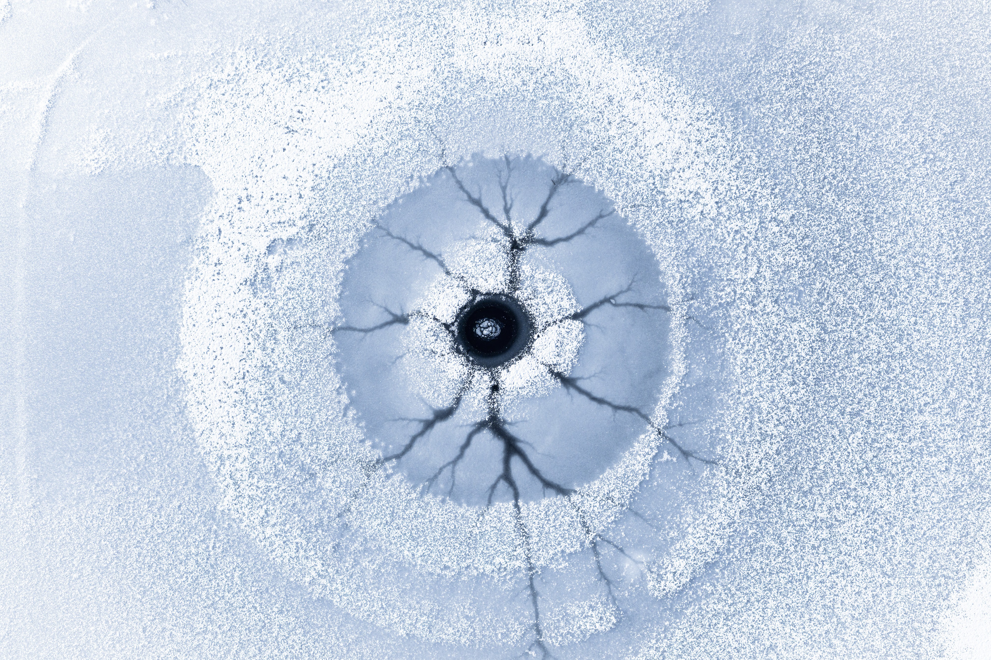 A top-down view of a circular opening in lake ice, with dark cracks radiating through the ice