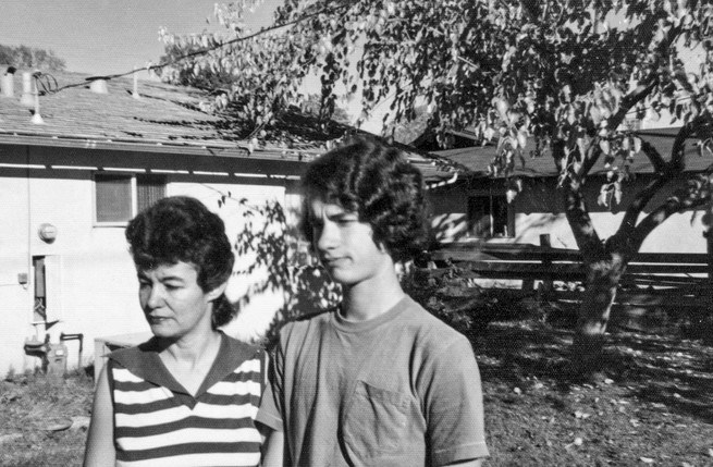 black-and-white candid photo of woman in striped shirt next to taller boy in yard with tree next to house