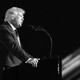 A black-and-white photograph of Donald Trump delivering a speech at a lectern, taken from the side