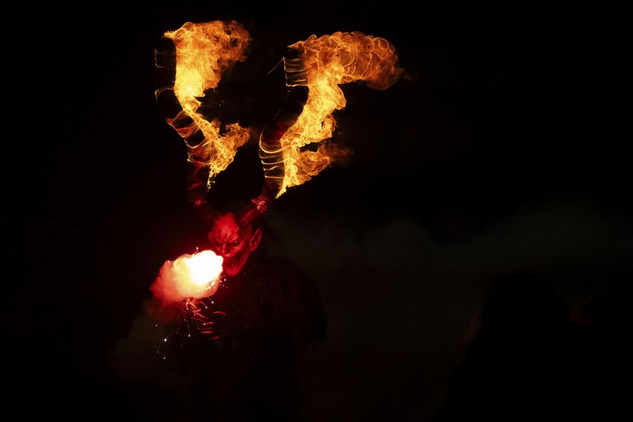 A person holds a torch while wearing a frightening demonic mask with long horns that are on fire