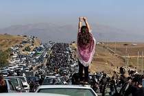 A woman holds her arms up, gesturing, while standing on a car among a large crowd of people marching past cars on a major road.