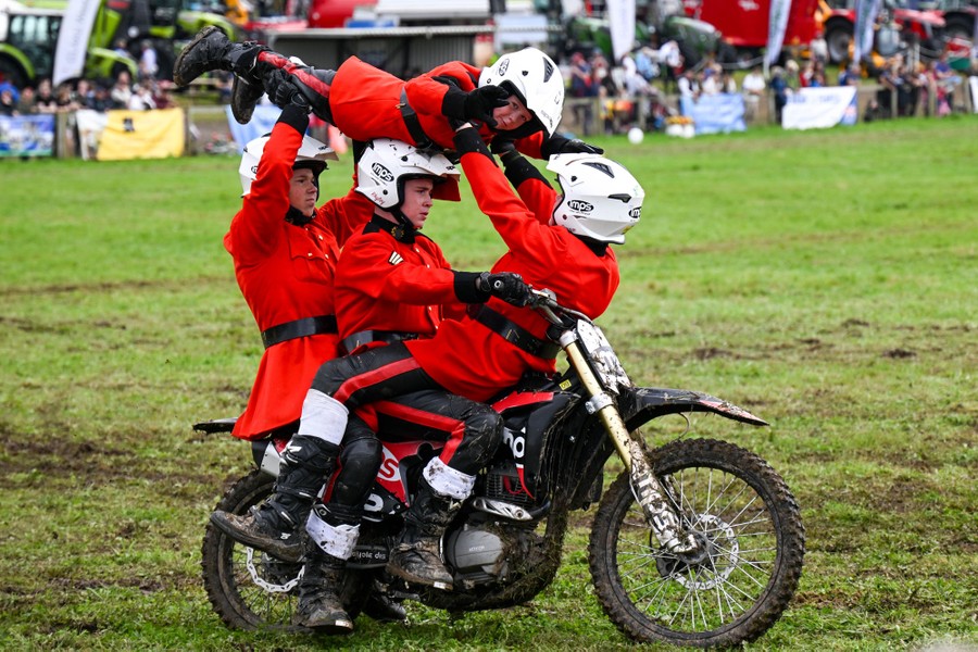 Four young uniformed motorcyclists arranged acrobatically ride a single motorcycle in a field.