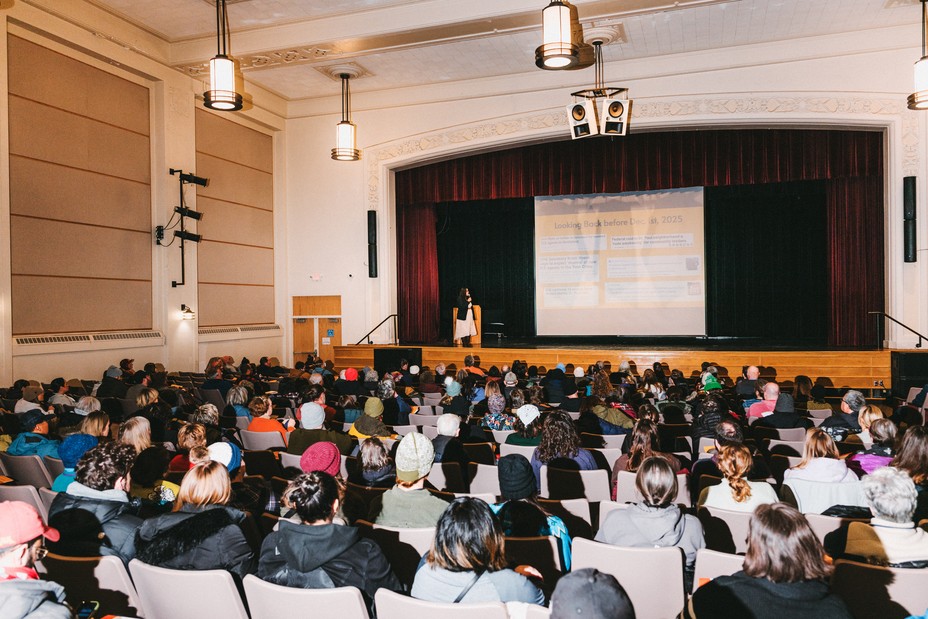 A person on a stage in an auditorium is seen in front of a large seated crowd