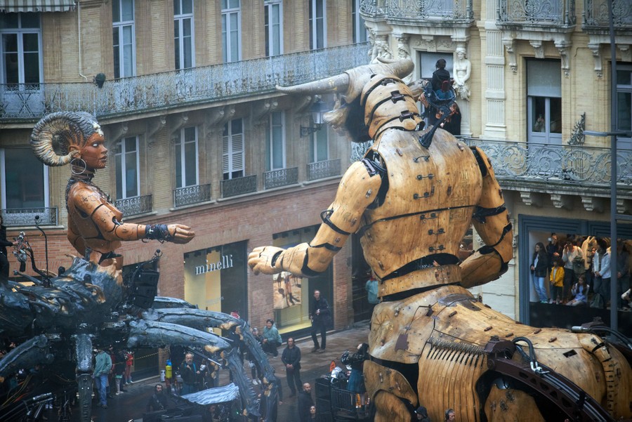 Two giant puppets interact alongside buildings.