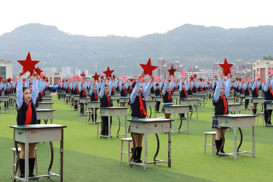 Young students stand beside many desks arranged in a grid on a playing field, each holding a red star above their heads.