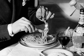 A black-and-white photograph of a torso in a suit and two hands swirling spaghetti against a spoon