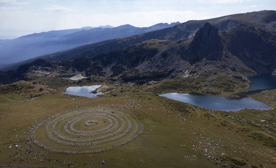 An elevated view of a large group of people wearing white clothing, standing in circles in a broad mountain valley.