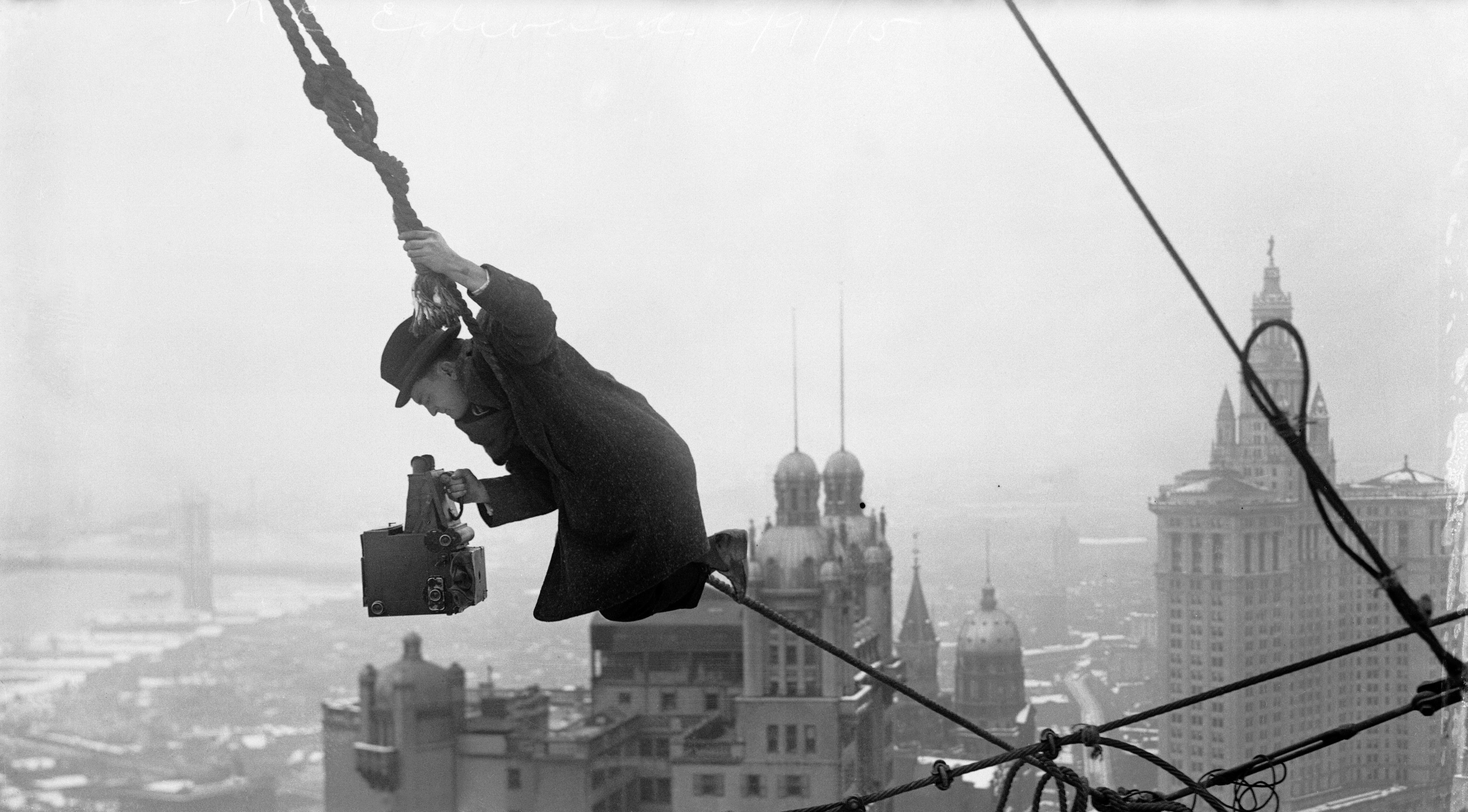 A photographer holding a large boxy camera hangs perilously from a rope on a high building, with a cityscape visible in the background.