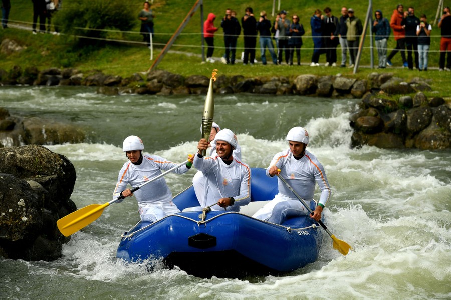 Several people row an inflatable raft down a whitewater course as one person rides in front, holding an Olympic torch aloft.