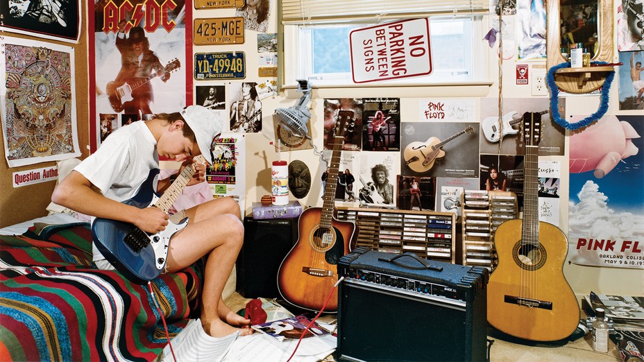 photo of boy sitting on bed playing electric guitar in room covered with posters, next to desk with racks of cassette tapes, an amp, and two acoustic guitars
