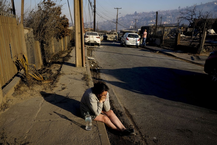 A person sits on a sidewalk near burned houses, crying.
