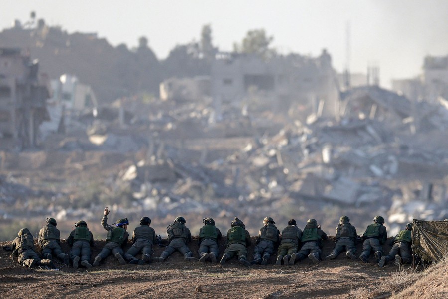 More than a dozen soldiers lie side-by-side along a ridge, looking out over a landscape filled with the rubble of destroyed buildings.