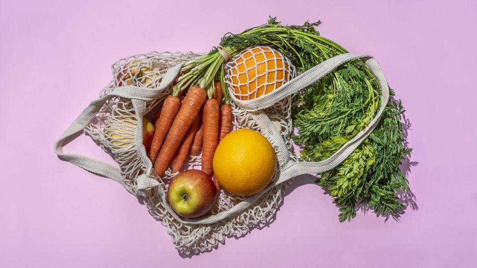 A woven bag containing carrots, apples, two oranges, and some greens, against a lavender background