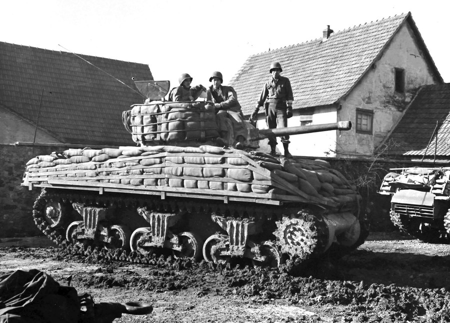 Three solders stand and sit atop an American tank that has been augmented with extra sandbags as armor.