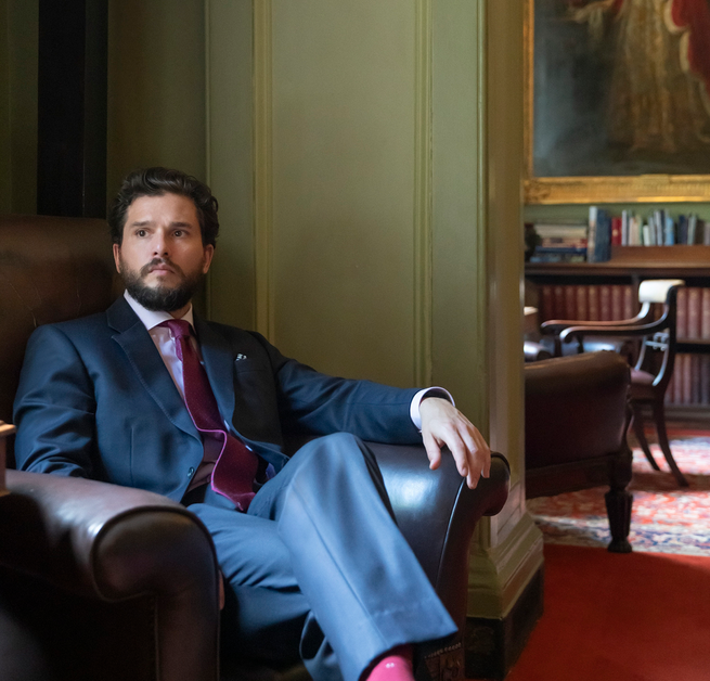 photo of bearded man in suit and tie sitting in large armchair in opulent room