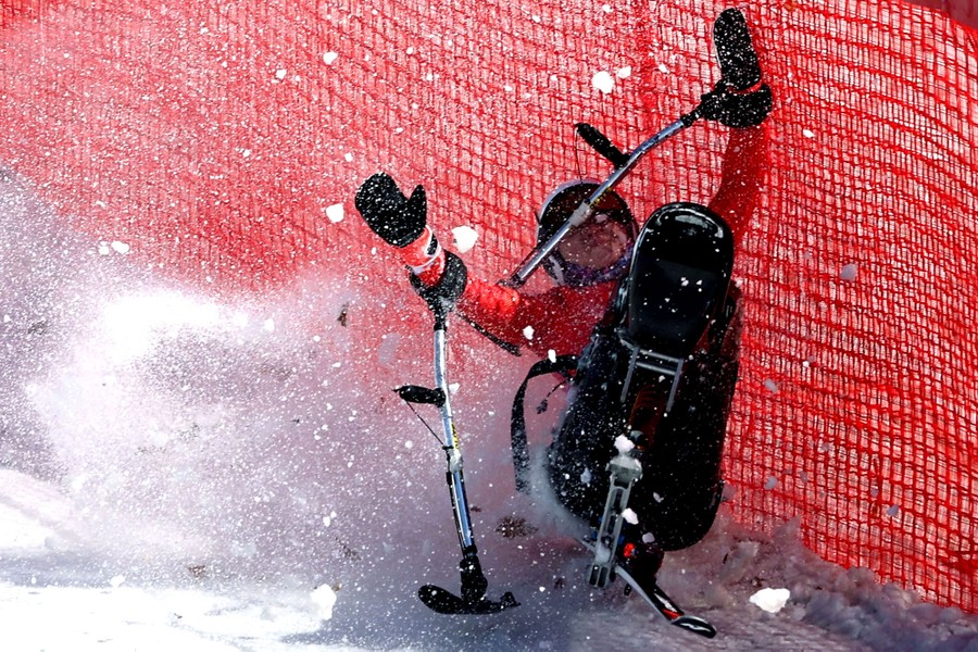 A seated skier crashes along a wall of netting, their arms held up, snow flying in the air.