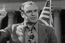 A man stands in front of an American flag with a pointed finger raised.