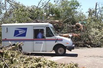 A U.S. Postal Service truck drives down a road lined by downed trees.