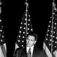 Black-and-white photo of Ron DeSantis speaking into a microphone, in front of three American flags