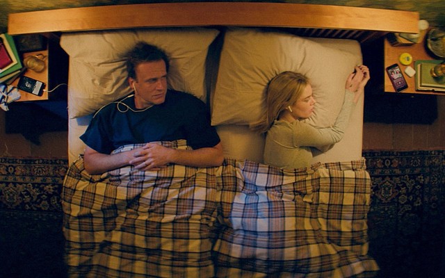 Jason Segel looks at Samara Weaving while lying in a bed with a plaid comforter