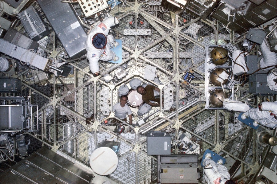 A view of the interior of a space station, with two men at center and lots of equipment and gear around them.