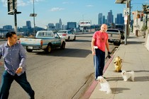 A man hurriedly walks away from another man, who is standing near a fire hydrant with two small white dogs.