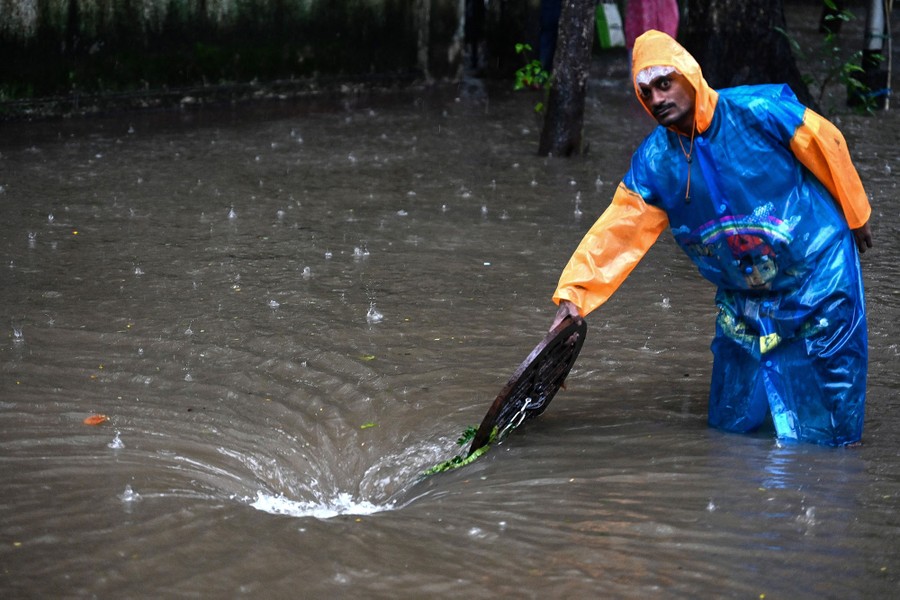 A person wearing a long raincoat stands in knee-deep floodwater beside an open manhole into which water is draining, forming a small whirlpool.