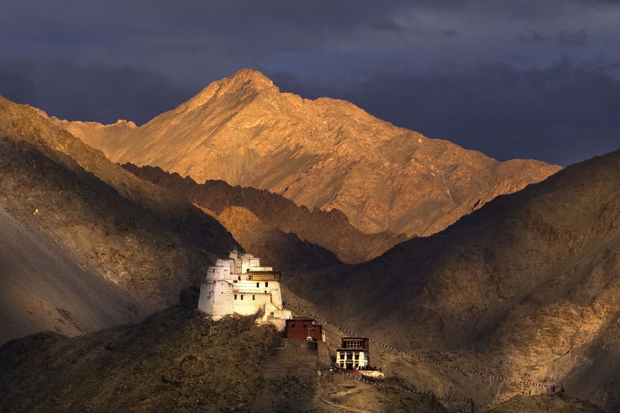 A monastery sits on a rocky mountaintop; larger mountains are visible in the background.