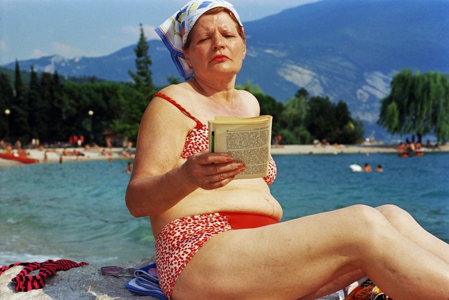 A woman in a kerchief and red bikini reads a book on a beach.