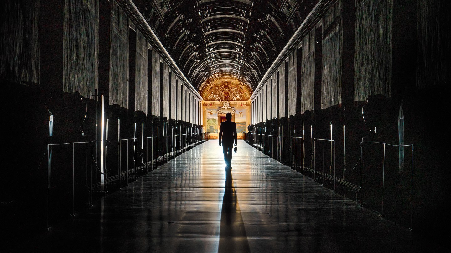Photo of a figure holding keys walking down a long, dark aisle toward a frescoed, lit door in the distance