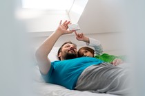 A father and son use an electronic device while lying in bed.