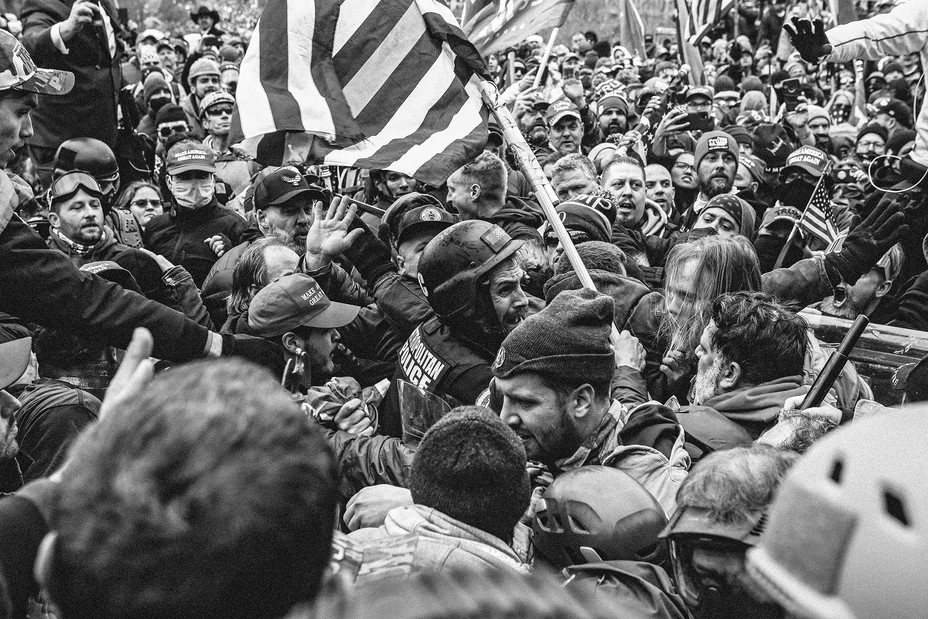 black and white photo of one police officer in helmet, face contorted, surrounded and confronted by enormous crowd, with one person brandishing an American flag