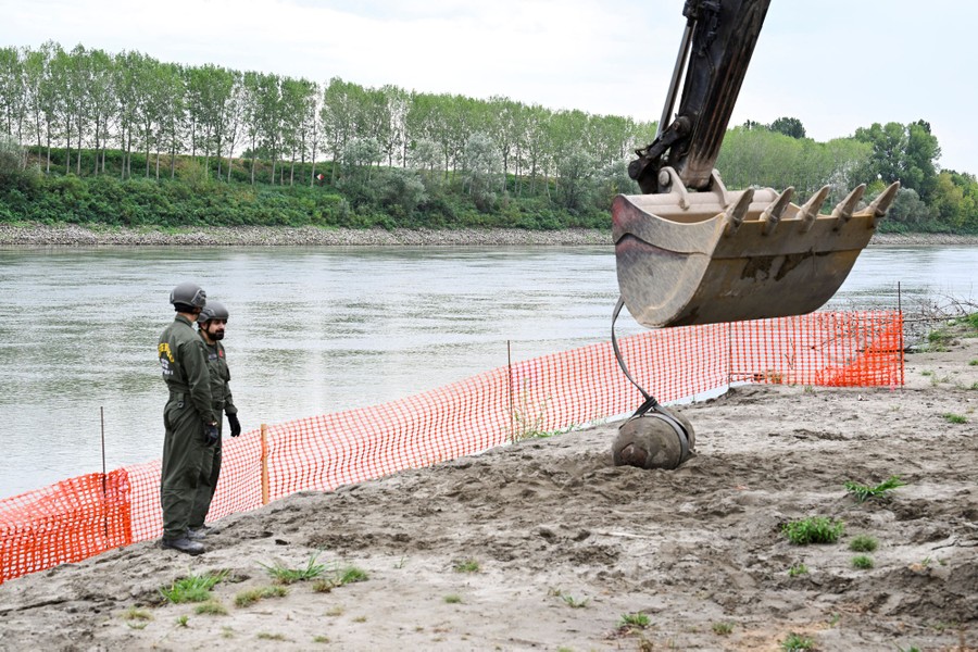 Two soldiers supervise as an excavator is used to lift a decades-old bomb from a riverbank.