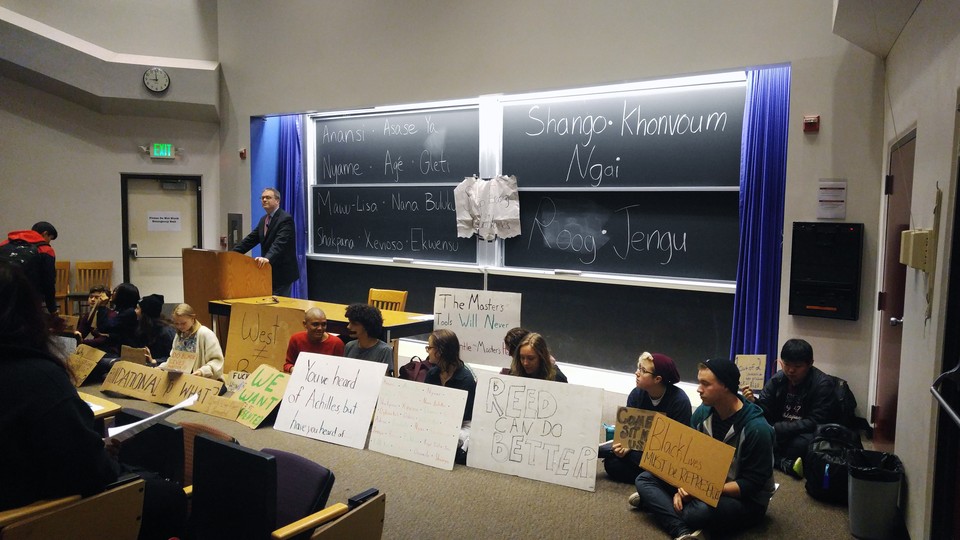 A professor stands at a lecture podium, while protesters sit alongside him holding signs. 
